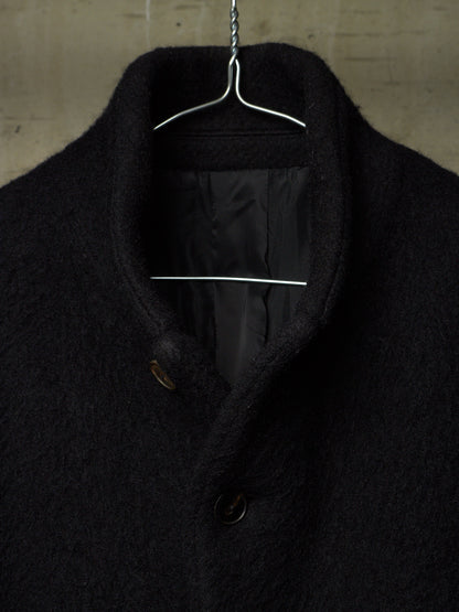A close-up of a black textured wool coat hanging on a metal hanger against a concrete wall.