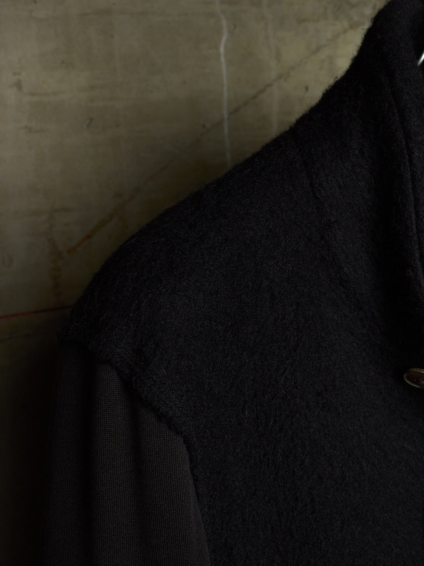 Close-up detail of a black textured wool-like garment, showing the shoulder and collar area against a neutral concrete-textured background.