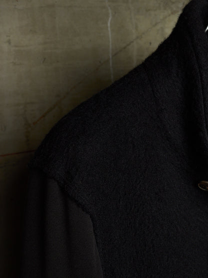 Close-up detail of a black textured wool-like garment, showing the shoulder and collar area against a neutral concrete-textured background.