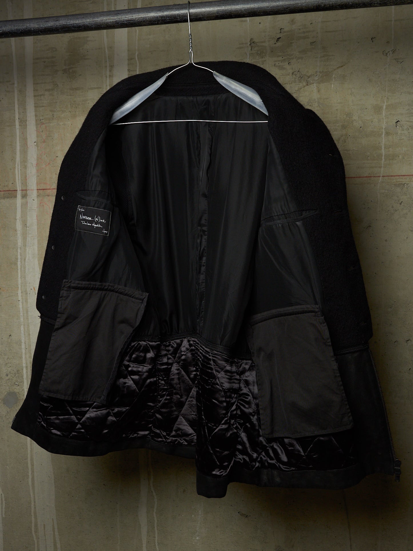 A black quilted jacket with a shiny black satin lining, hanging on a metal hanger against a textured concrete wall.