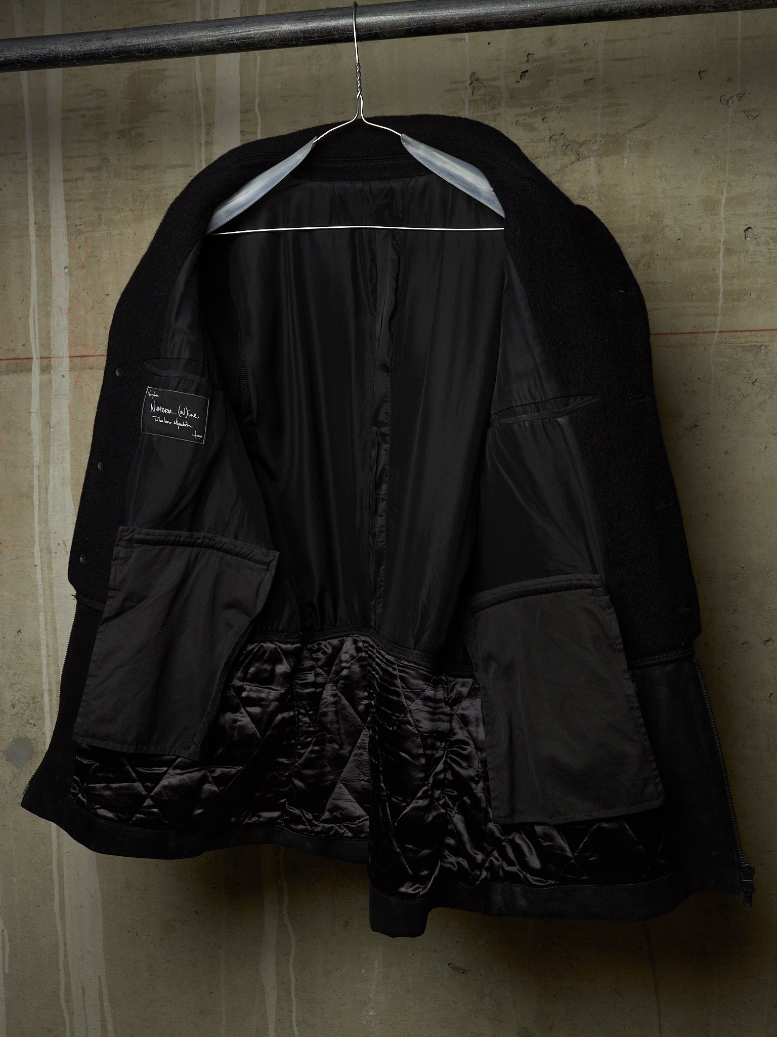 A black quilted jacket with a shiny black satin lining, hanging on a metal hanger against a textured concrete wall.