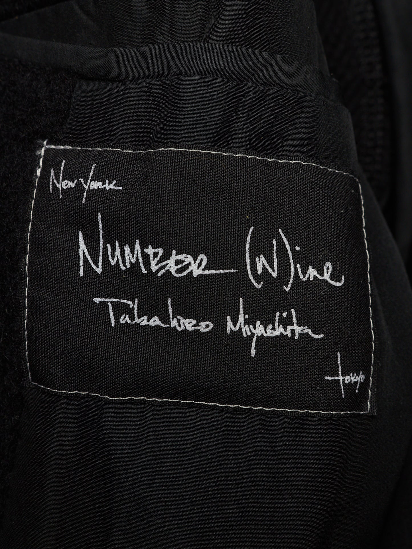 Close-up of a black fabric patch with white stitched border and white handwritten text reading 'Number (W)ine' and 'Takeshi Nojima', attached to a black garment.