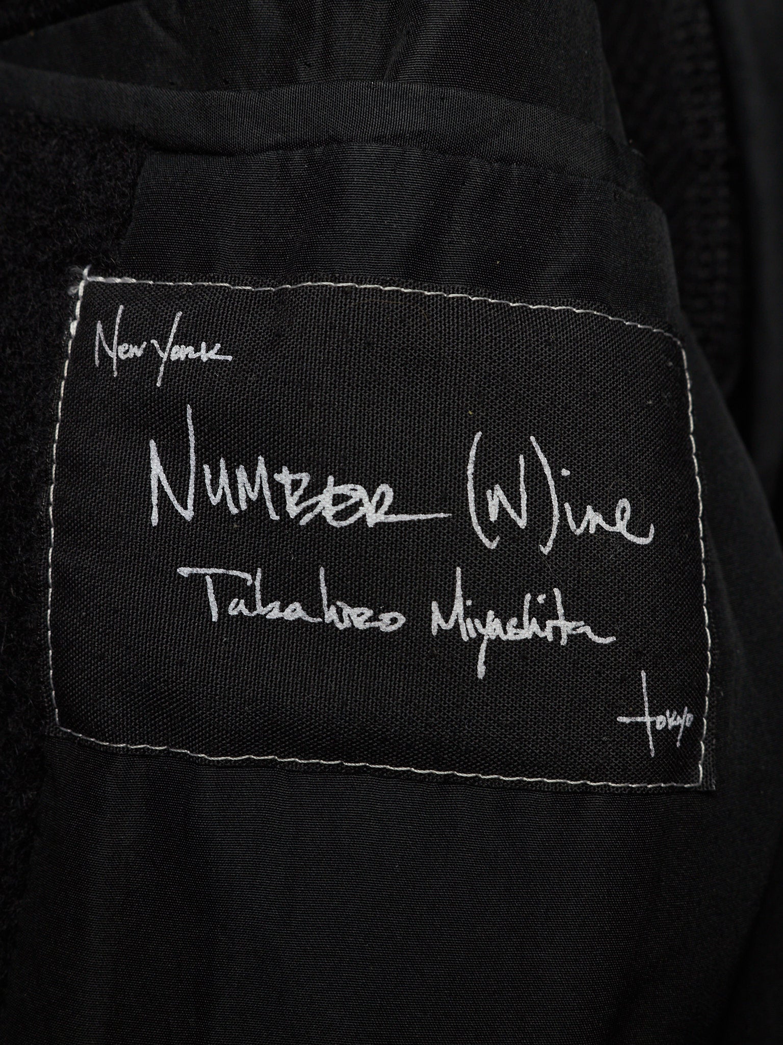 Close-up of a black fabric patch with white stitched border and white handwritten text reading 'Number (W)ine' and 'Takeshi Nojima', attached to a black garment.