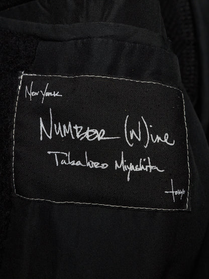 Close-up of a black fabric patch with white stitched border and white handwritten text reading 'Number (W)ine' and 'Takeshi Nojima', attached to a black garment.