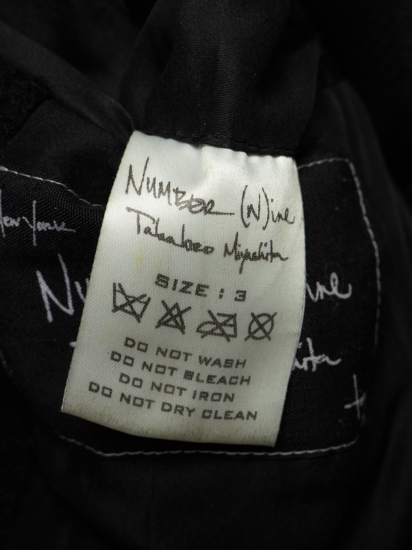 A close-up of a black garment's interior label, featuring a white rectangular tag with handwritten size information and care instructions. The handwritten text identifies the brand as 'Number (Wine) Takahiro Miyakita'.