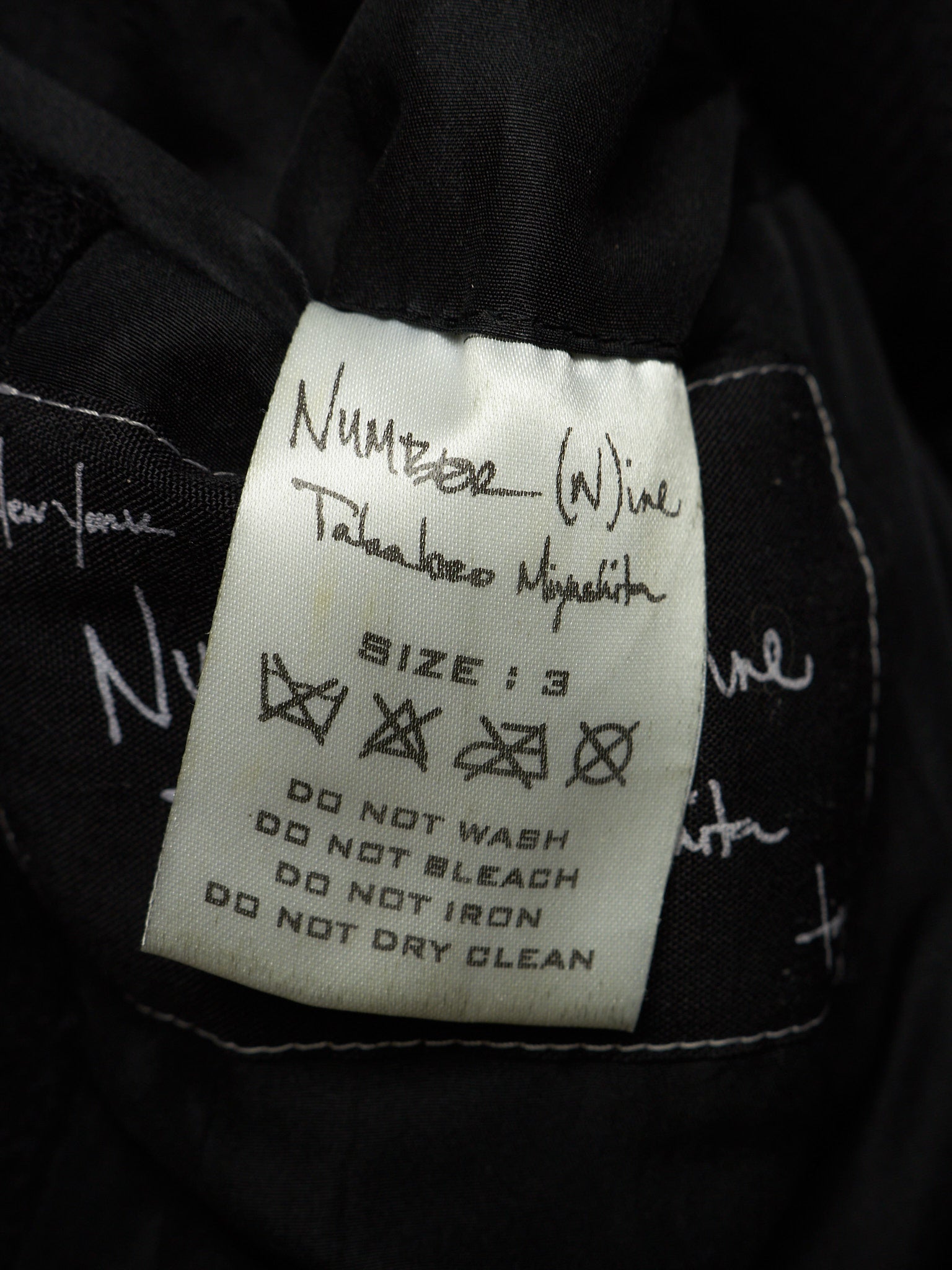A close-up of a black garment's interior label, featuring a white rectangular tag with handwritten size information and care instructions. The handwritten text identifies the brand as 'Number (Wine) Takahiro Miyakita'.