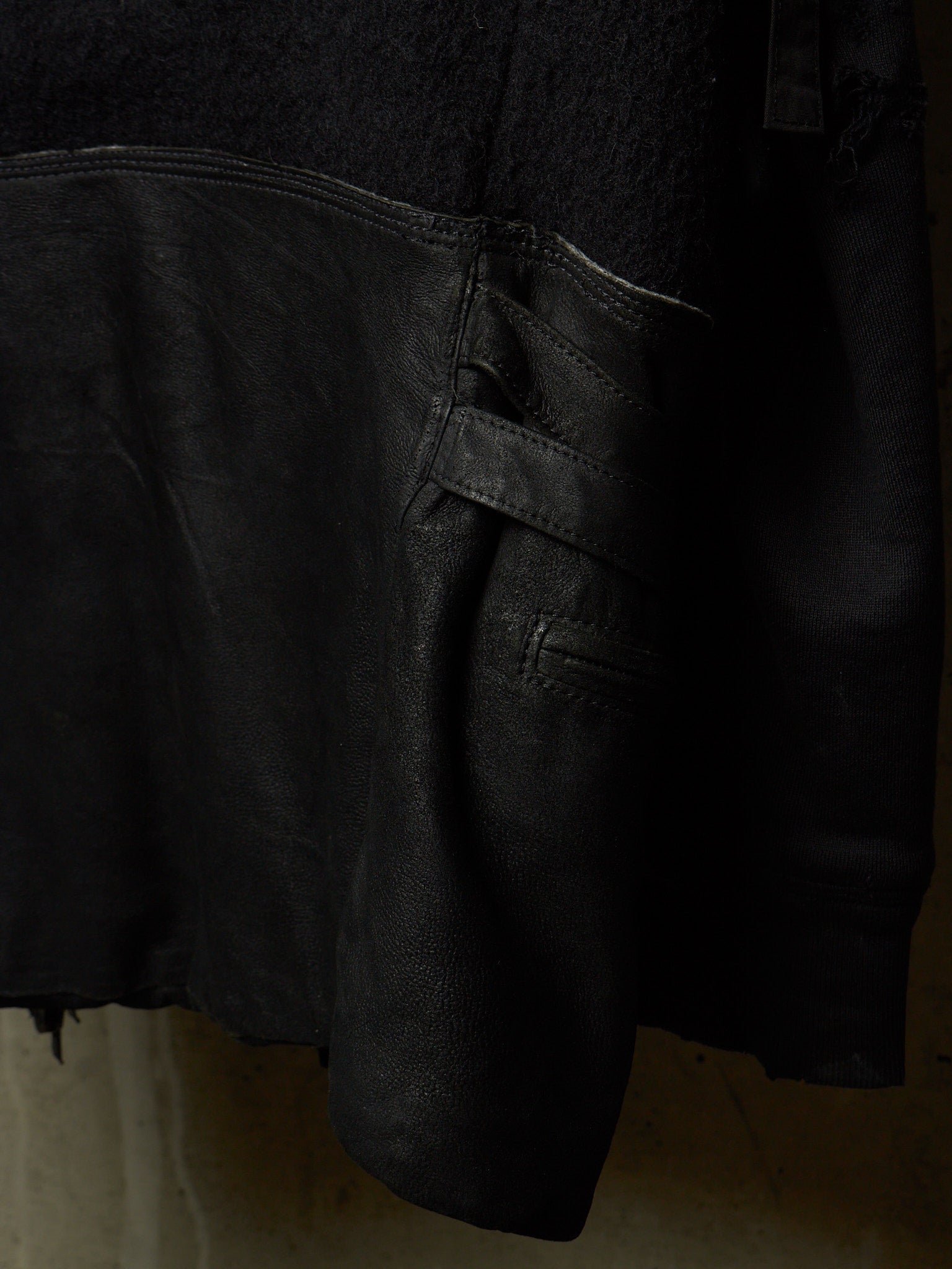Close-up detail of a black leather jacket showing the texture of the leather, a side pocket with a strap closure, and a contrasting black fabric section at the top.