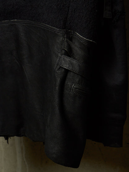 Close-up detail of a black leather jacket showing the texture of the leather, a side pocket with a strap closure, and a contrasting black fabric section at the top.