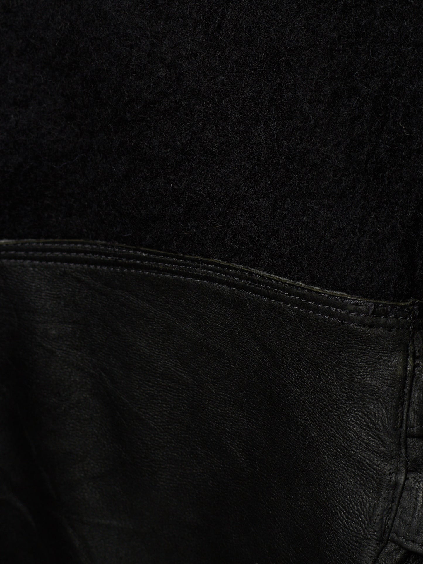 Close-up detail of a black garment showing a textured black fabric upper section and a black leather lower section with visible stitching.