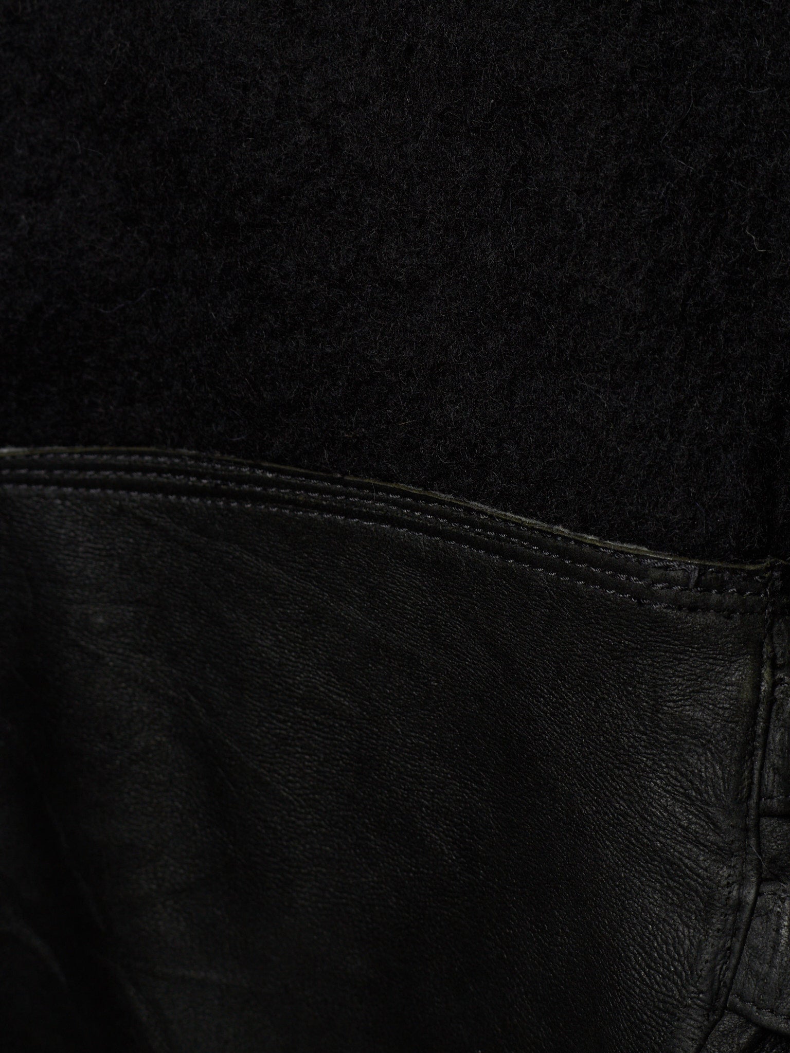 Close-up detail of a black garment showing a textured black fabric upper section and a black leather lower section with visible stitching.