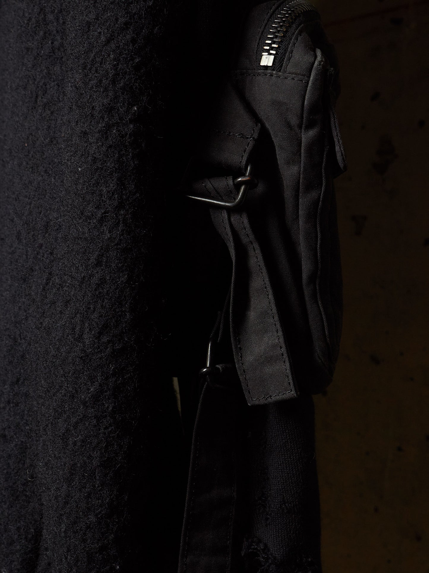 A close-up of a black crossbody bag with a textured fabric body, black strap, and silver-toned zipper, shown against a dark background.