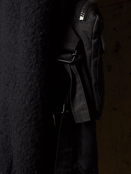 A close-up of a black crossbody bag with a textured fabric body, black strap, and silver-toned zipper, shown against a dark background.