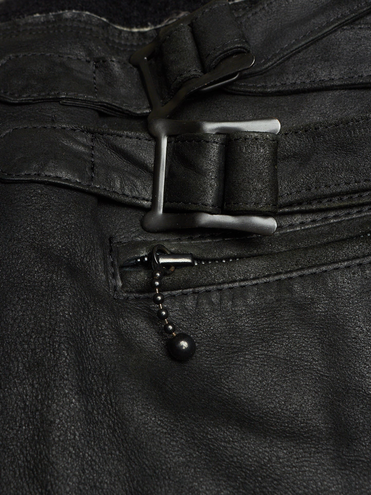 Close-up of black leather pants showing a belt buckle, belt strap, zipper pocket, and a small black bead chain detail.