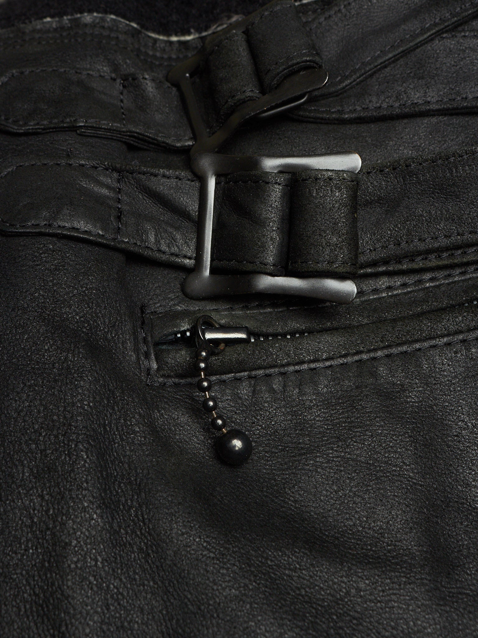 Close-up of black leather pants showing a belt buckle, belt strap, zipper pocket, and a small black bead chain detail.