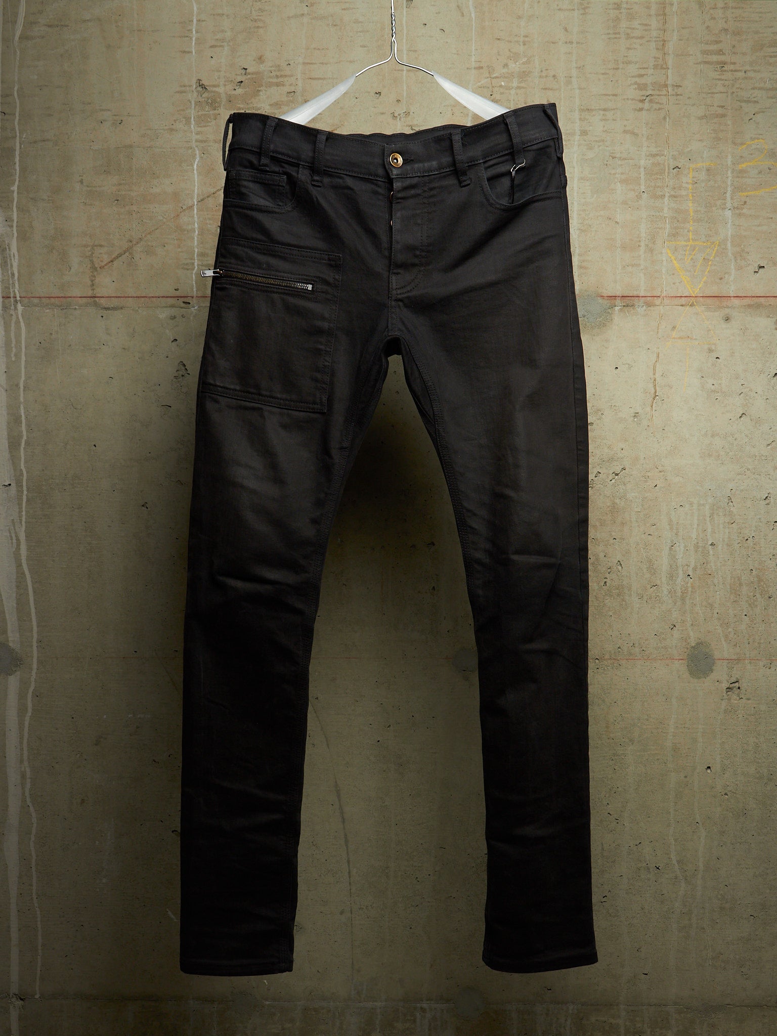Black slim-fit jeans with zippered side pockets, hanging on a white wire hanger against a textured concrete wall.