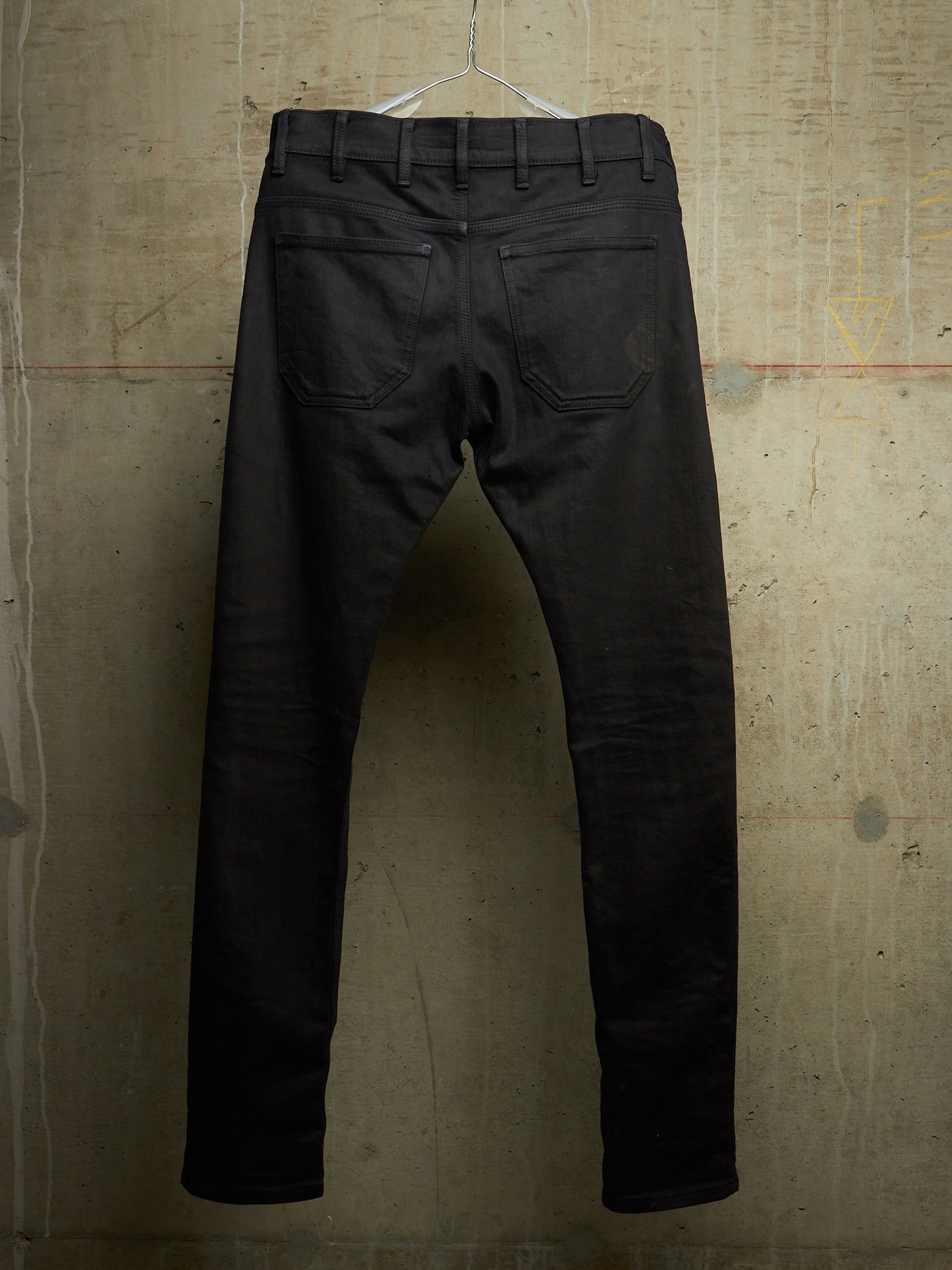 Black slim-fit jeans shown from the back, hanging on a clear plastic hanger against a textured concrete wall.