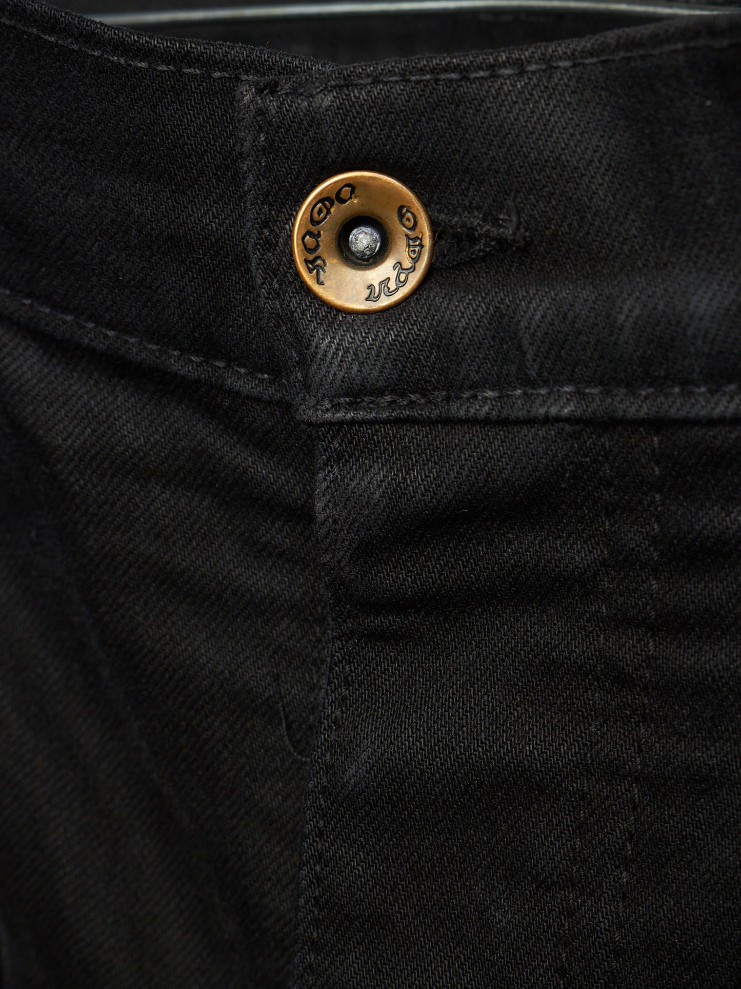 Close-up of black denim jeans showing the waistband and a round brass button with engraved branding.