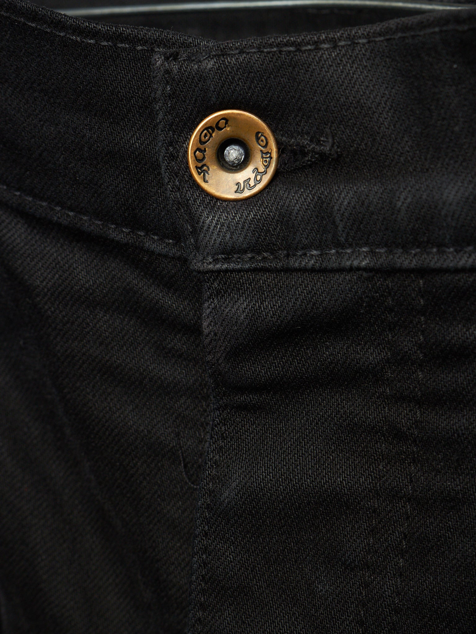 Close-up of black denim jeans showing the waistband and a round brass button with engraved branding.