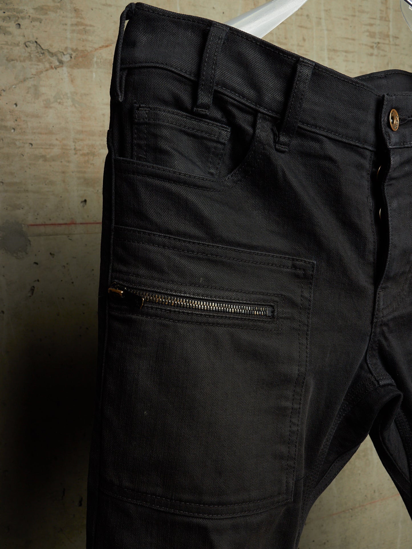 Close-up of black denim jeans hanging against a worn beige floor, showing the back pocket area with a horizontal zip pocket and belt loops.