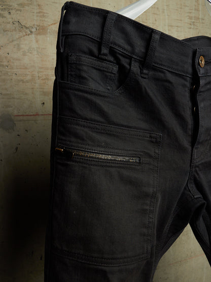 Close-up of black denim jeans hanging against a worn beige floor, showing the back pocket area with a horizontal zip pocket and belt loops.