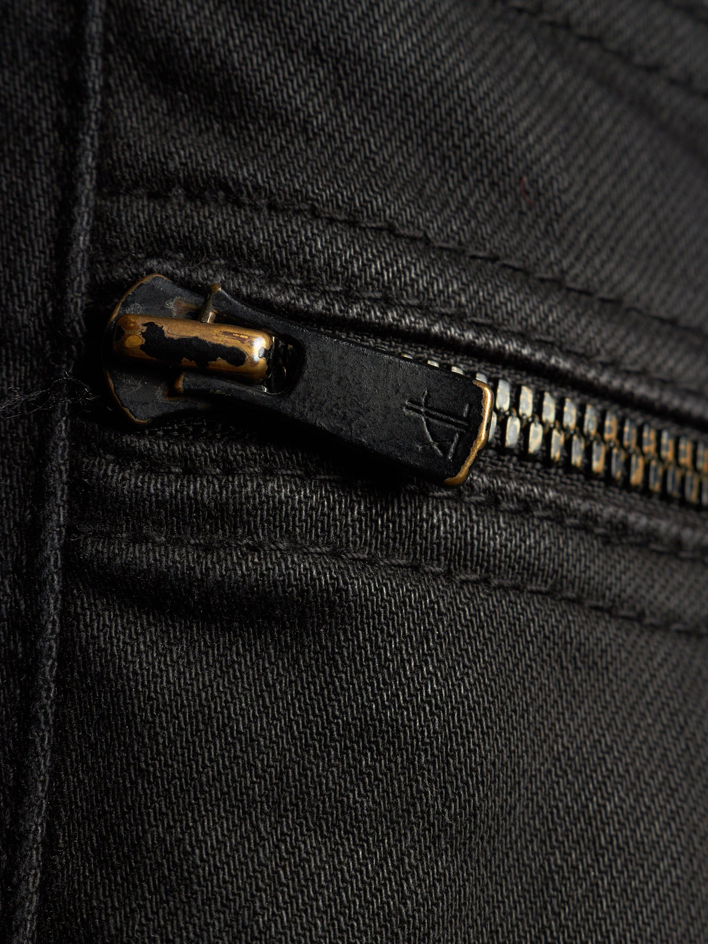 Close-up of a black denim or canvas zipper pocket with a black pull tab and brass-toned zipper teeth.