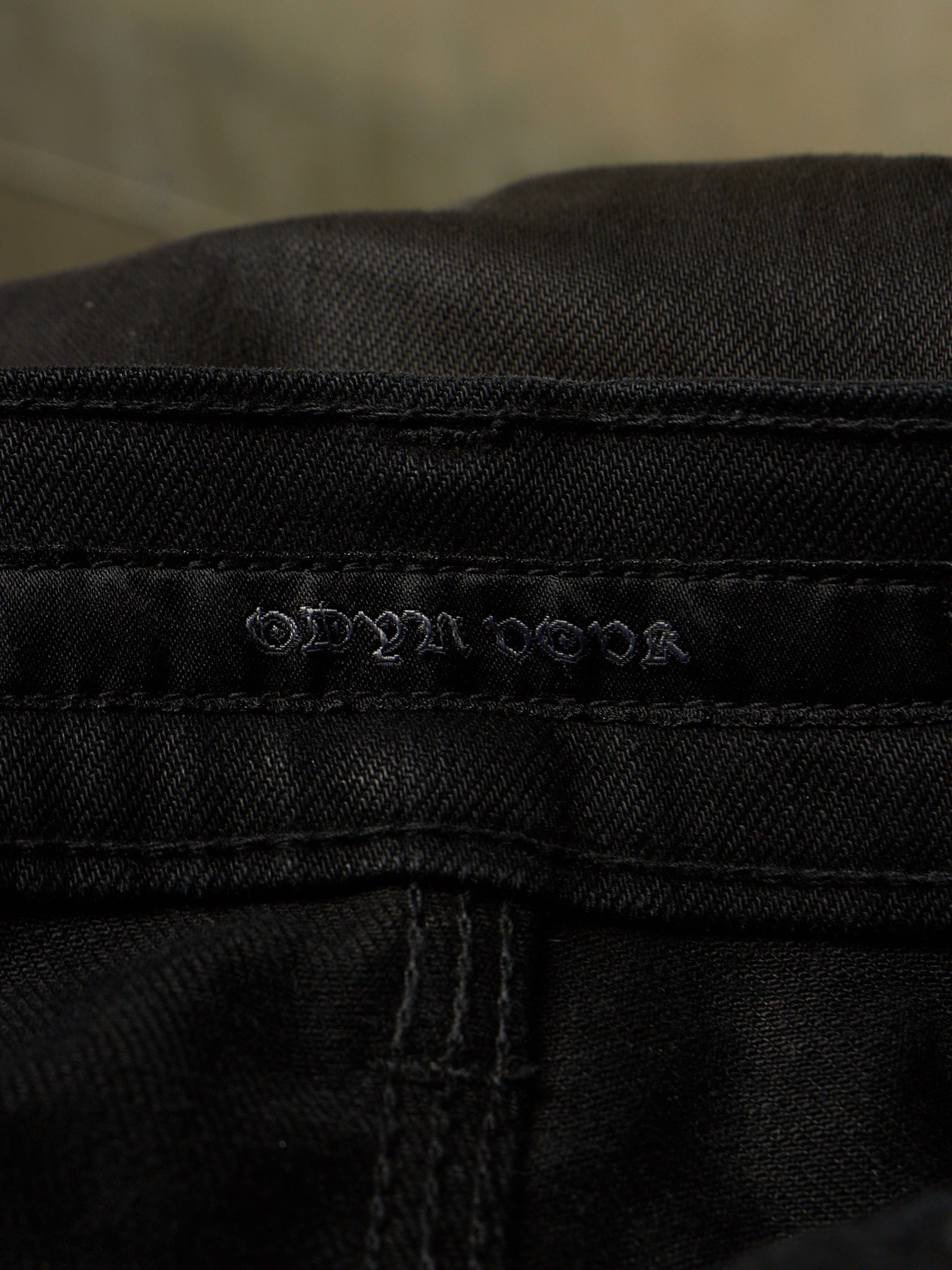 Close-up of black denim fabric showing the embossed brand name 'ODYSSEUS' on the waistband.