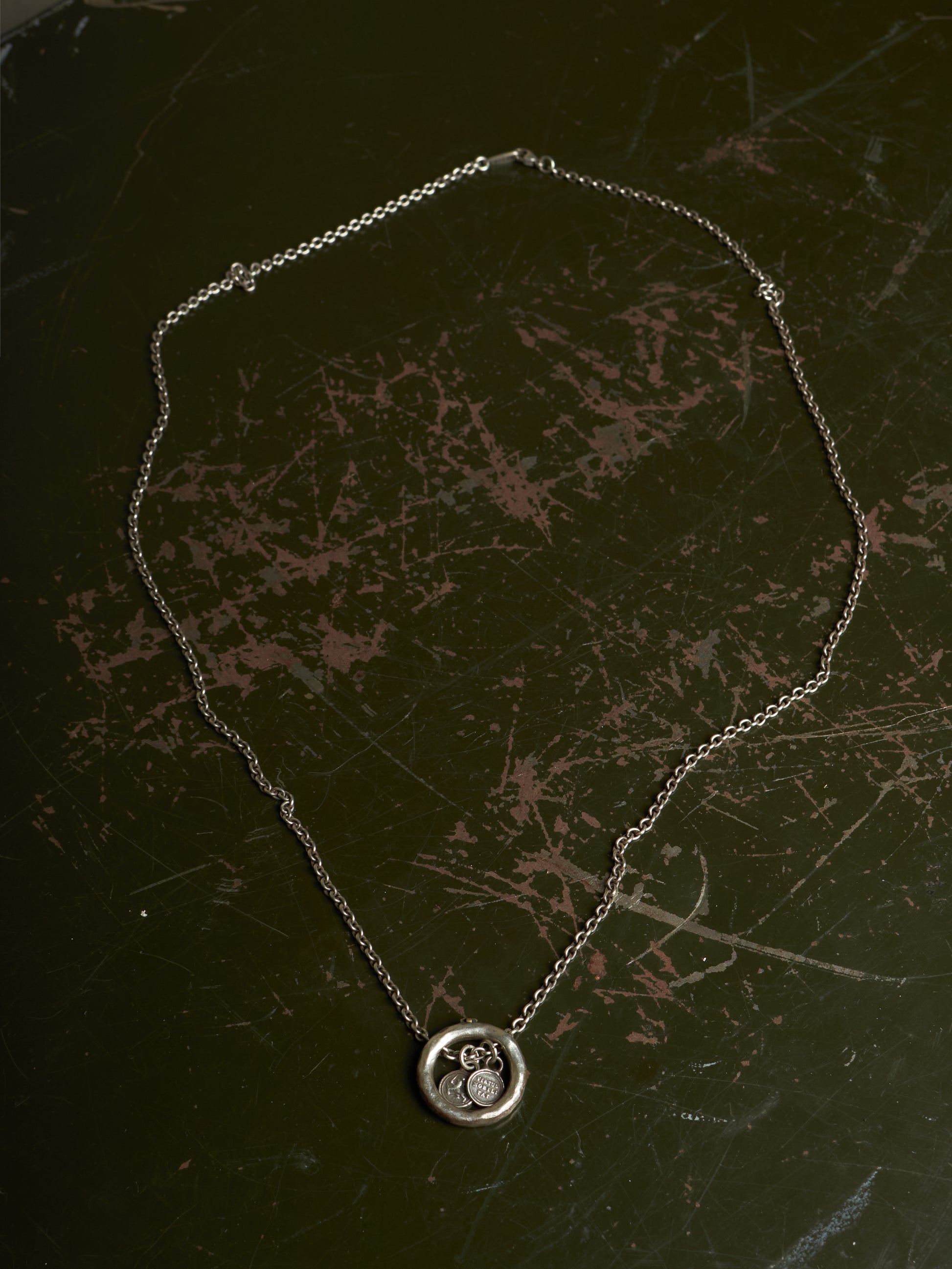A silver chain necklace with a circular pendant featuring a stylized cat design, displayed on a dark, scratched surface.