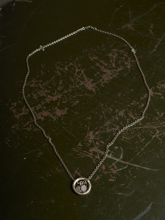 A silver chain necklace with a circular pendant featuring a stylized cat design, displayed on a dark, scratched surface.
