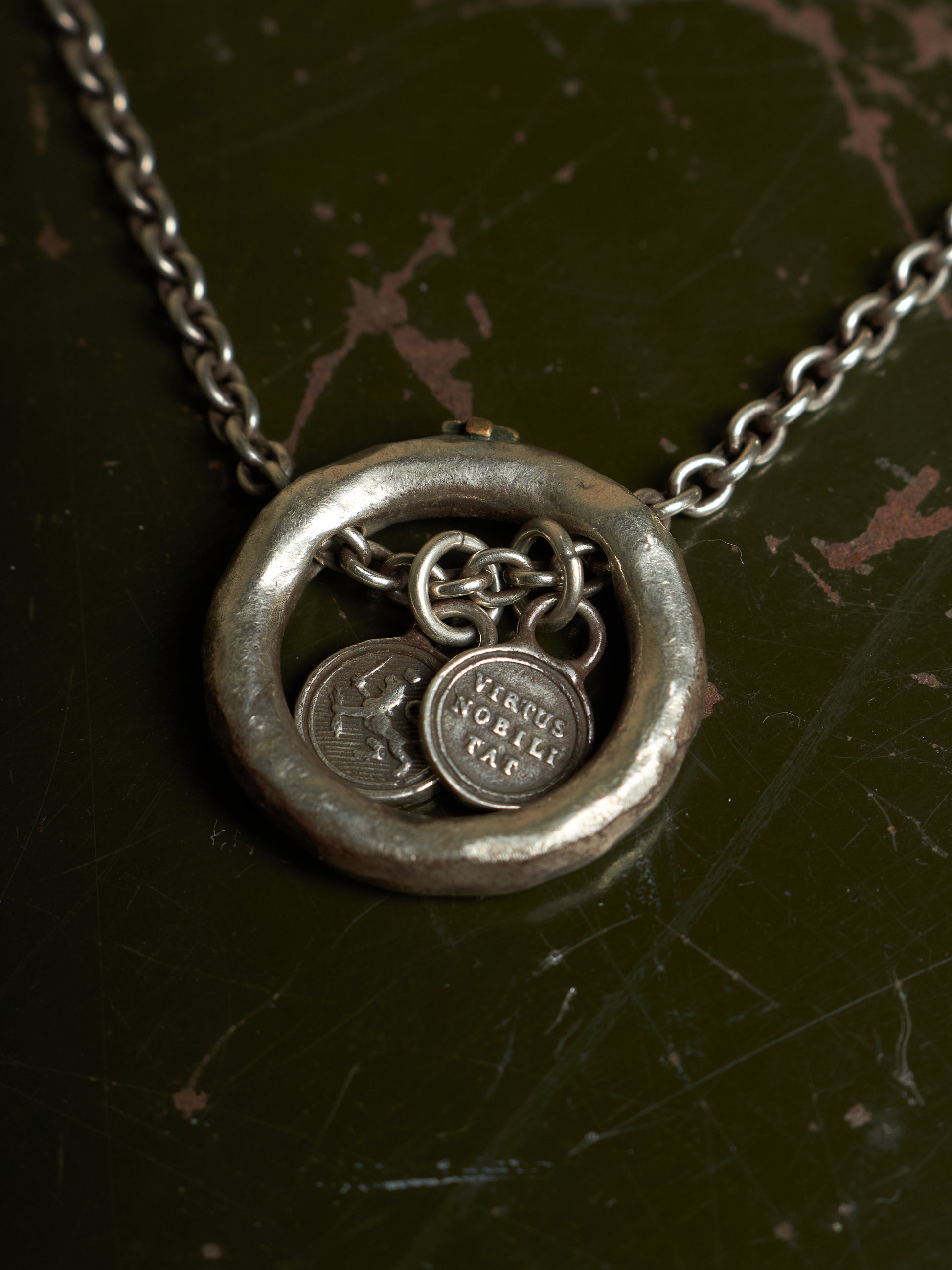 A silver-toned necklace with a textured circular pendant and dangling coin charms, displayed on a dark, distressed background.