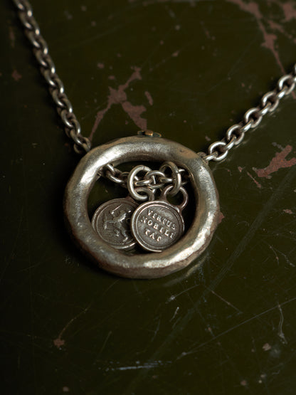 A silver-toned necklace with a textured circular pendant and dangling coin charms, displayed on a dark, distressed background.