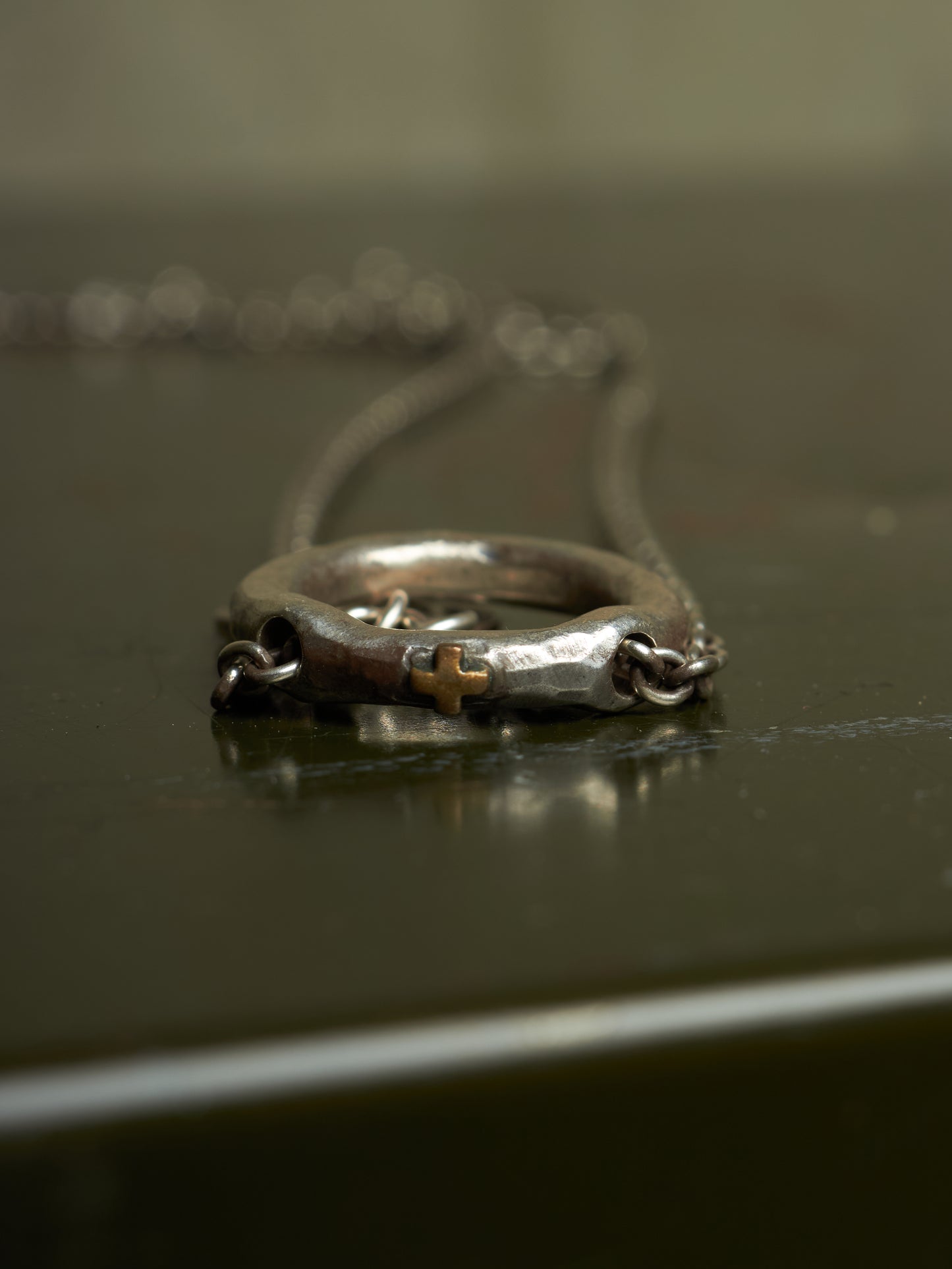 A close-up of a silver-toned ring necklace with a small gold cross detail, resting on a dark reflective surface.