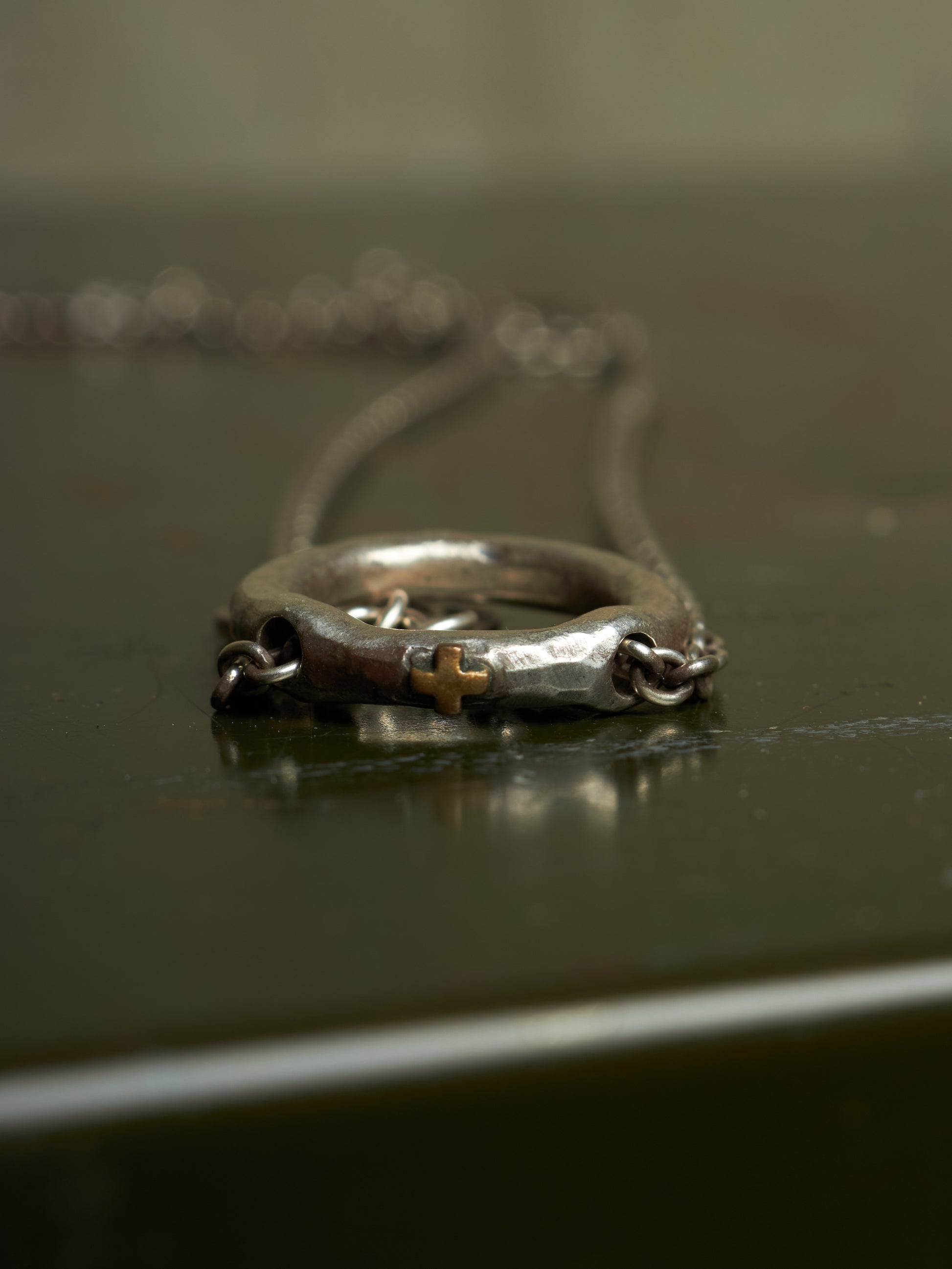A close-up of a silver-toned ring necklace with a small gold cross detail, resting on a dark reflective surface.