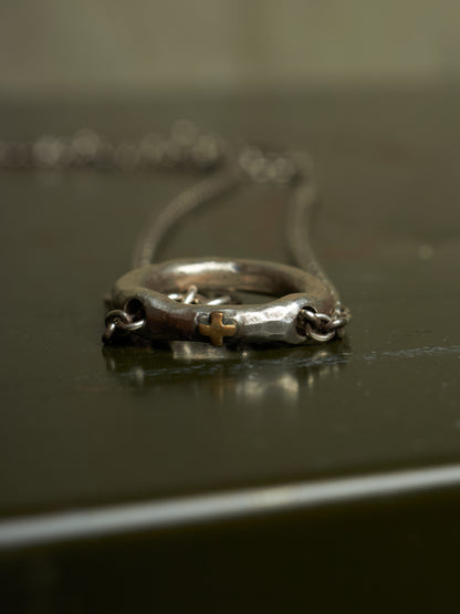 A close-up of a silver-toned ring necklace with a small gold cross detail, resting on a dark reflective surface.