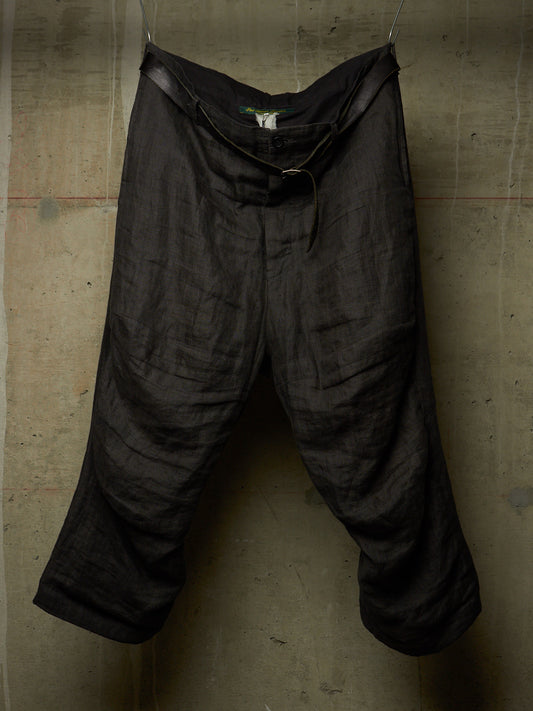 Black linen trousers with a drawstring waist, hanging against a textured concrete wall.