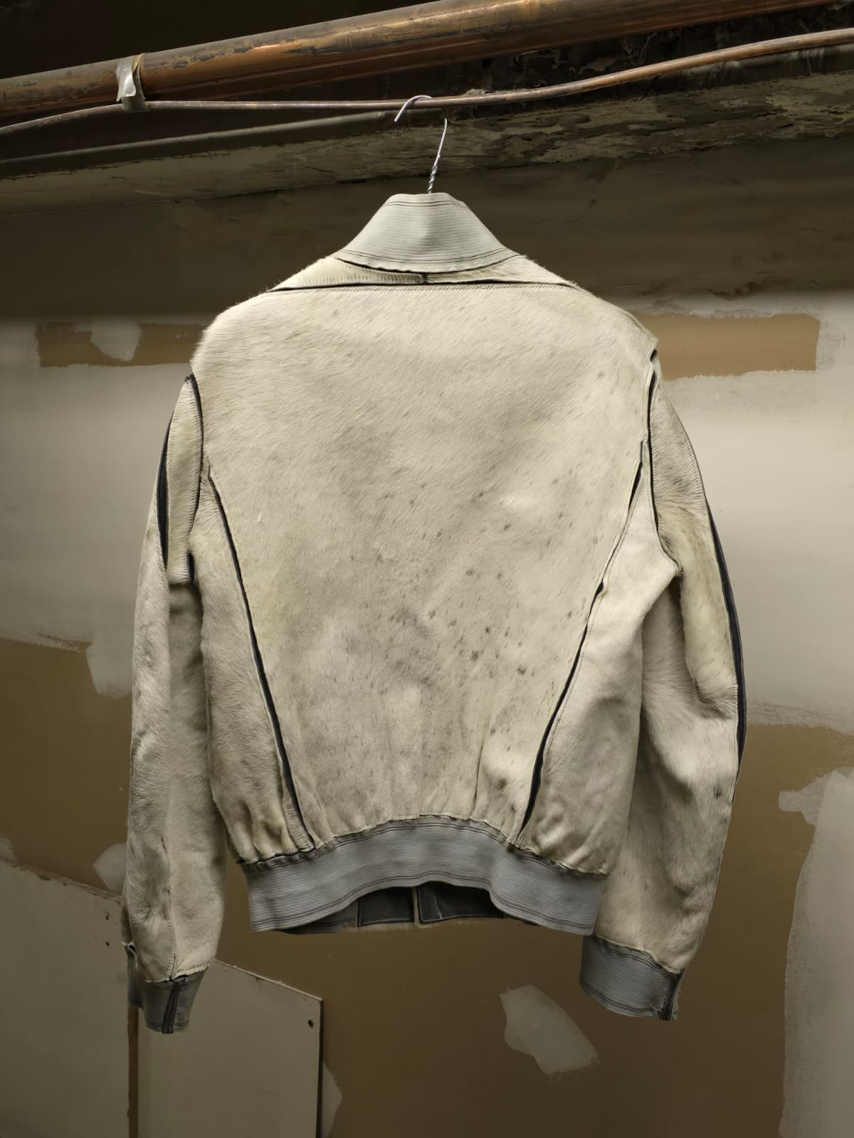 A light beige leather jacket with black contrast stitching, shown from the back and hanging on a wire hanger against a plaster wall.