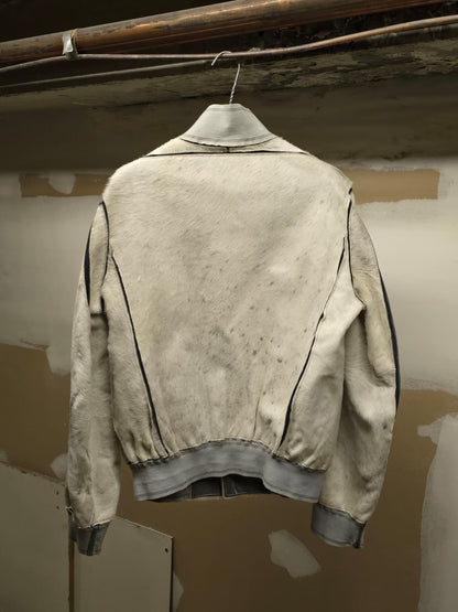 A light beige leather jacket with black contrast stitching, shown from the back and hanging on a wire hanger against a plaster wall.