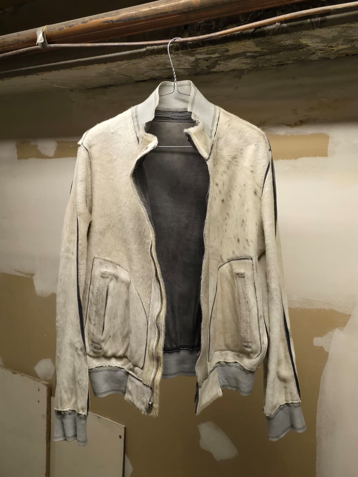 A distressed off-white leather jacket with gray ribbed trim, hanging on a wire hanger against a textured plaster wall.