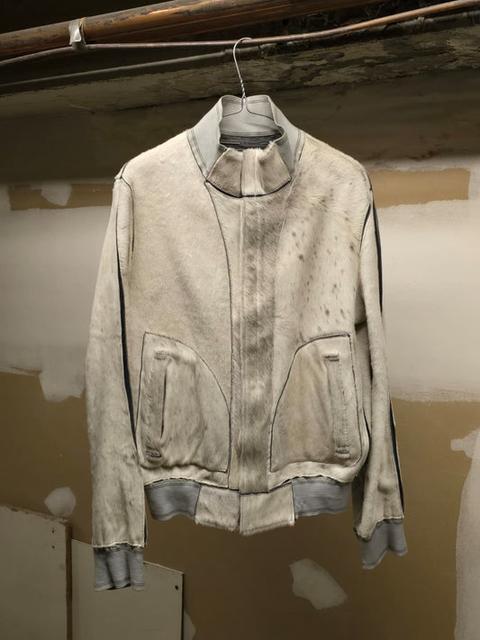 A distressed, light gray weathered jacket with a high collar and front zipper, hanging on a wire hanger against a plaster wall.