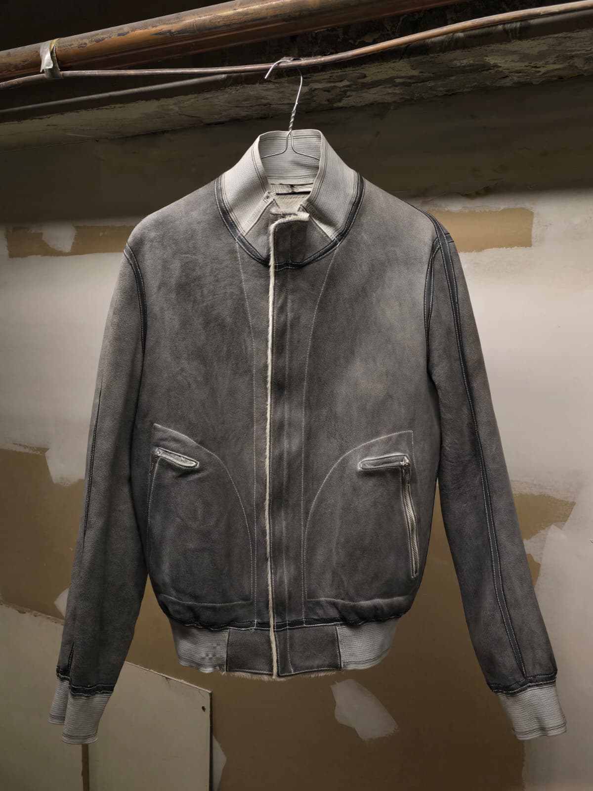 A dark gray washed suede bomber jacket with a stand collar and front zipper, hanging on a wire hanger against a rustic plaster wall.
