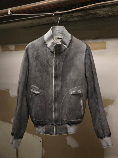 A dark gray washed suede bomber jacket with a stand collar and front zipper, hanging on a wire hanger against a rustic plaster wall.