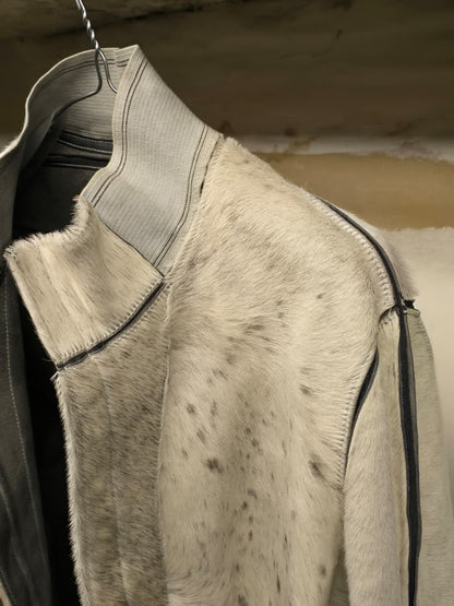 A close-up of a light gray and white speckled fur jacket with black trim, hanging on a metal hanger against a rustic wooden background.