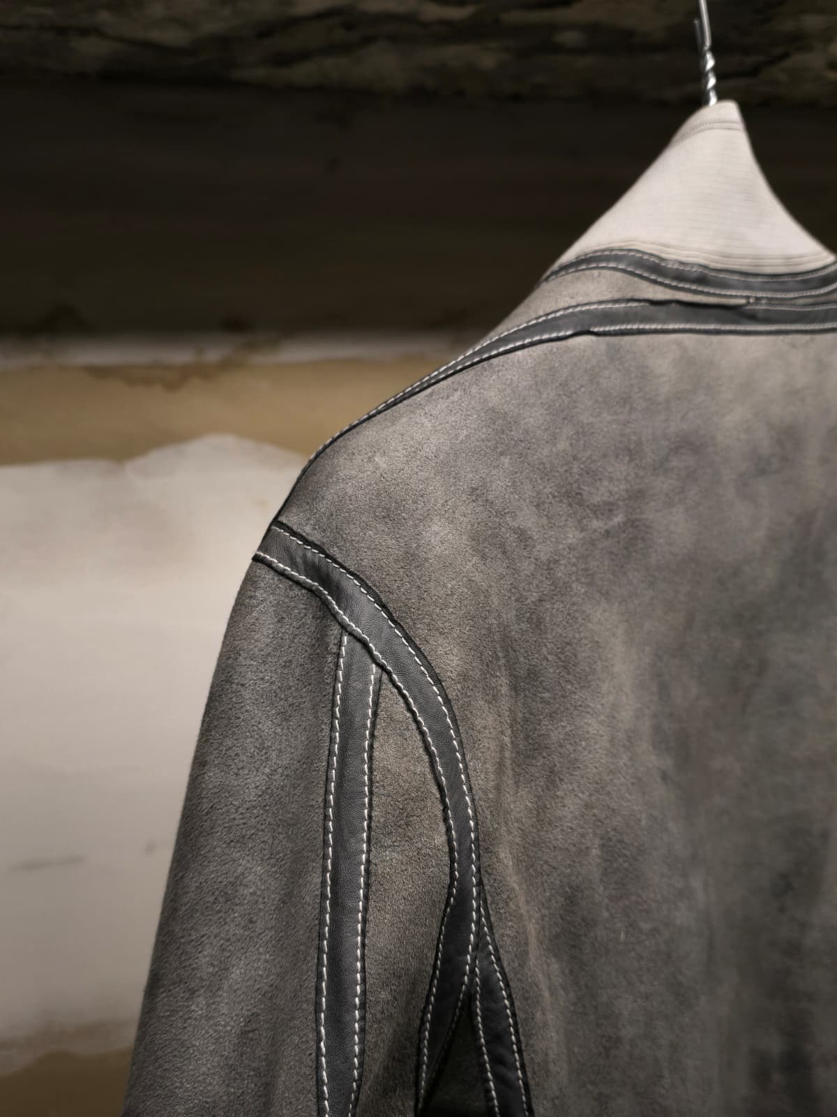 Close-up of a dark gray suede or nubuck leather jacket hanging on a white hanger, showing the textured fabric and contrast stitching.