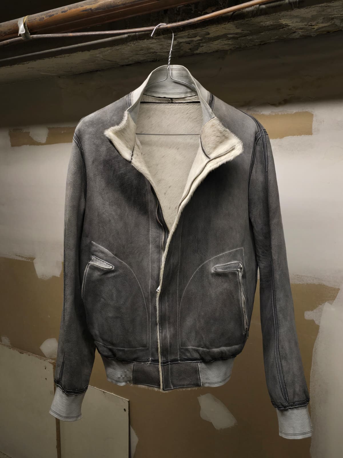 A dark gray suede bomber jacket with a cream-colored shearling lining, hanging on a wire hanger against a rustic, unfinished plaster wall.