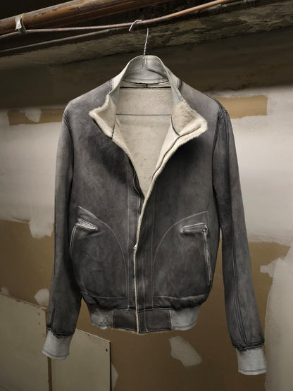 A dark gray suede bomber jacket with a cream-colored shearling lining, hanging on a wire hanger against a rustic, unfinished plaster wall.