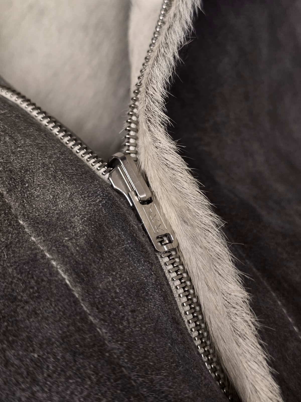 Close-up of a dark gray suede or shearling-lined jacket with a silver metal zipper and a light gray faux-fur trim.