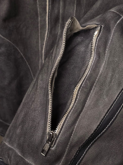 Close-up of a dark gray suede or nubuck jacket with a silver metal zipper pocket, showing the texture of the material and the contrast stitching.