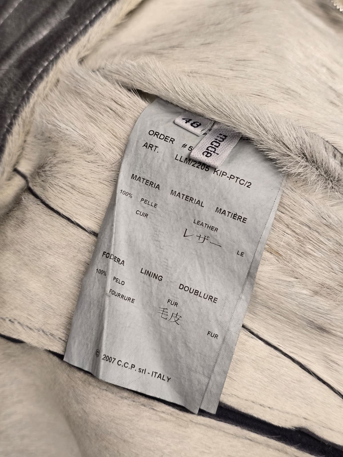 A close-up of a light gray fur garment with a white paper size tag sewn inside near the collar, showing the brand C.C.