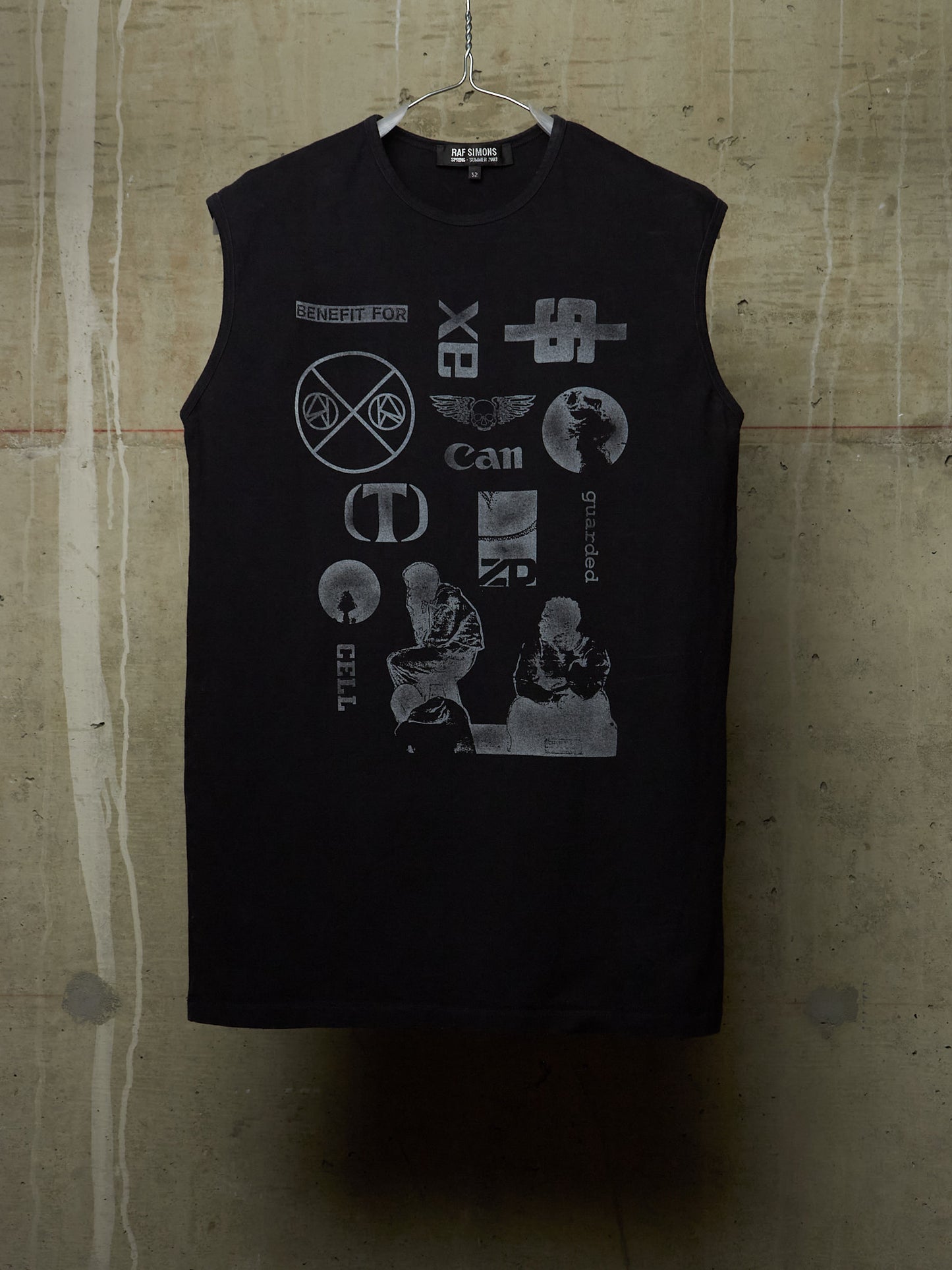 A black sleeveless tank top hanging on a wire hanger against a textured concrete wall. The shirt features a large, distressed gray graphic print on the front with various band logos and text, including 'Cult', 'Can', 'Sex', and 'Bitch'.