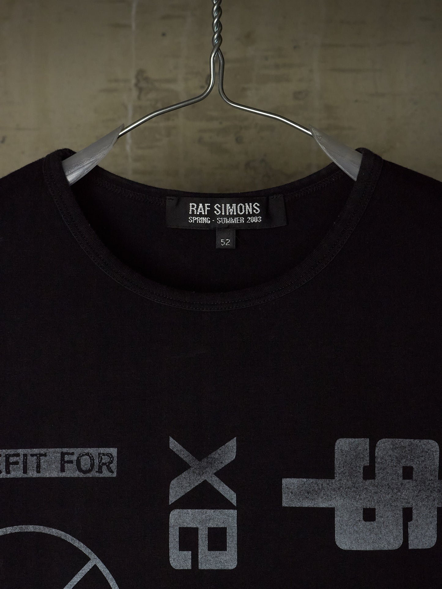 A black Raf Simons t-shirt hanging on a metal hanger against a textured concrete wall. The shirt features a gray graphic print on the front and a visible Raf Simons brand label inside the collar.