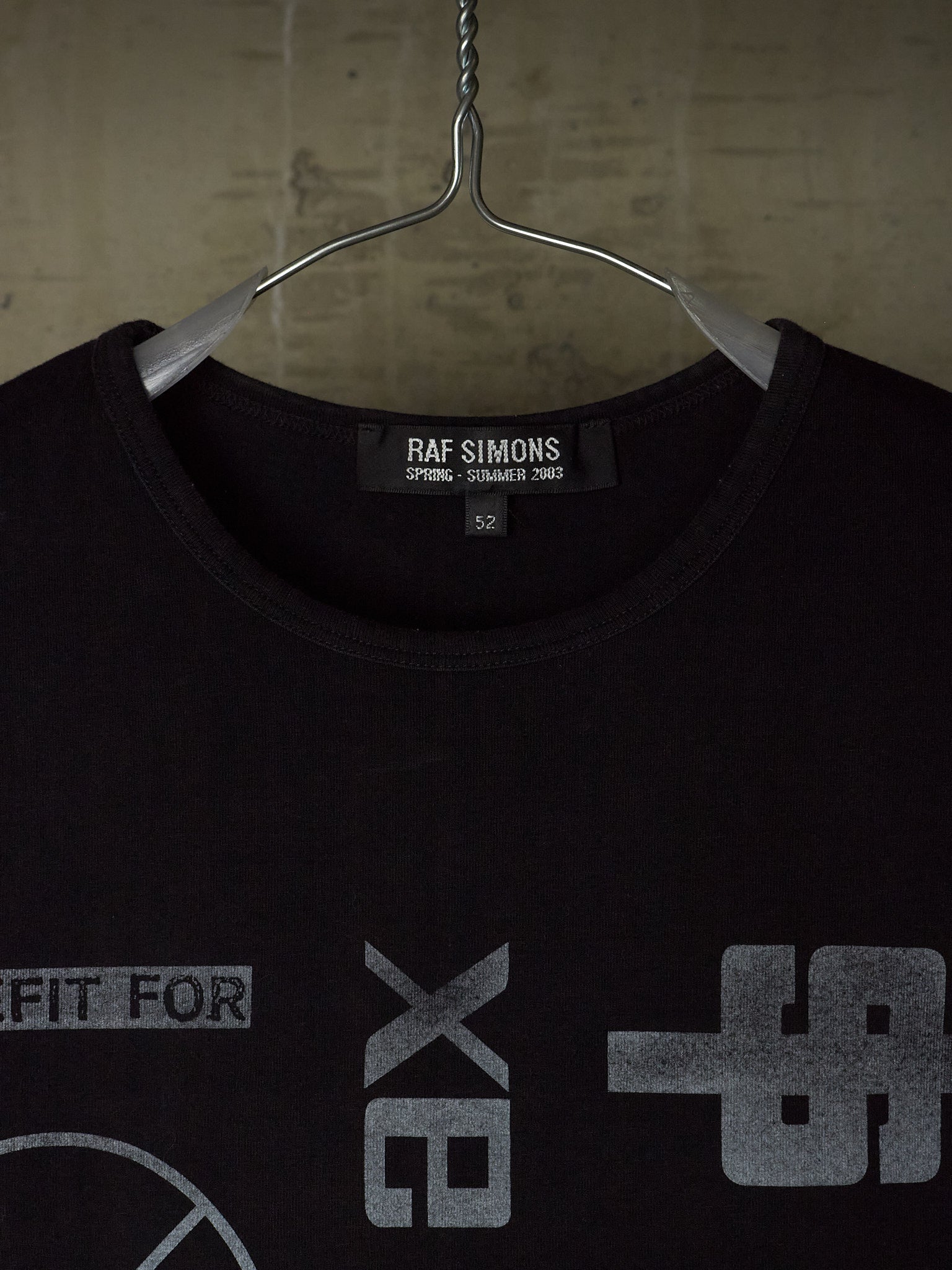 A black Raf Simons t-shirt hanging on a metal hanger against a textured concrete wall. The shirt features a gray graphic print on the front and a visible Raf Simons brand label inside the collar.